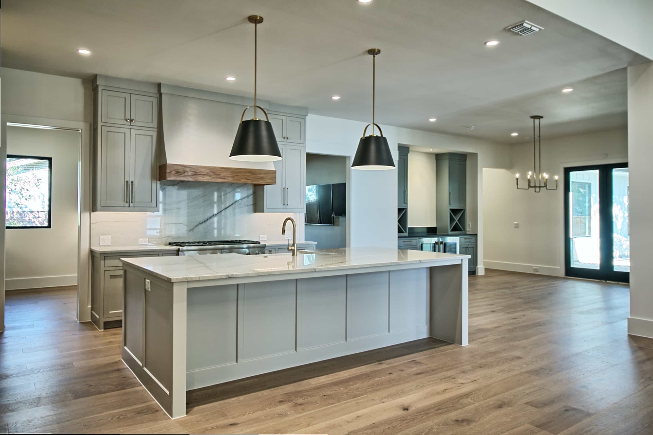 Kitchen with gray shaker cabinets, wood-banded hood, veined slab backsplash, large island with brass faucet and black pendants, open to dining area
